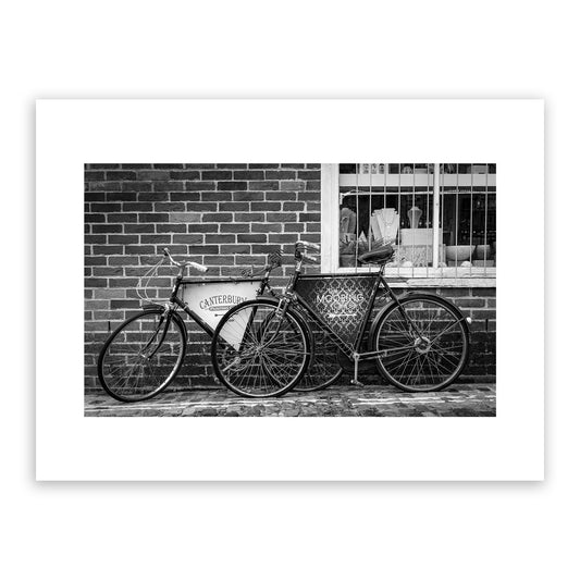 Old vintage bikes on the charming, cobbled, Water Lane, Canterbury, Kent, England, with signs for Canterbury Punting Company and The Mooring Café Bar. Rock, Paper, Scissors store visible through window. Fine art black and white photography print for sale.